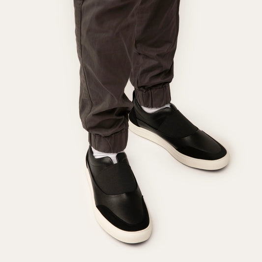 Black slip-on shoes worn with dark pants on a white background